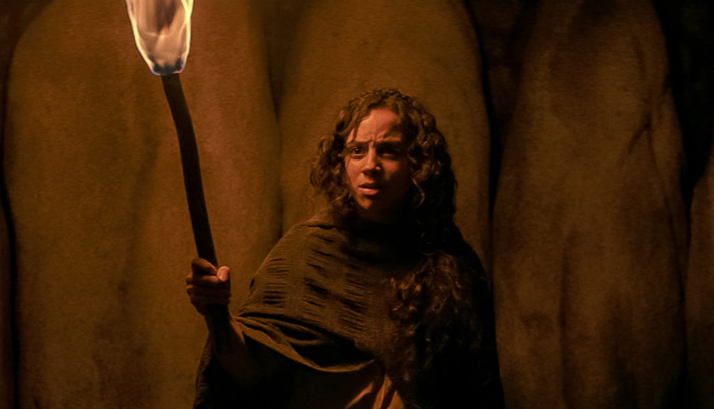 A person in a dark space holds a torch aloft and looks quite concerned.